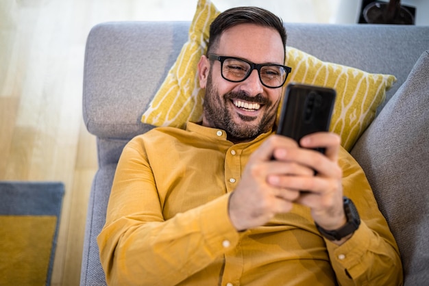 Allegro giovane uomo maturo adulto felice sorridente utilizzando smart phone che riposa sdraiato sul divano