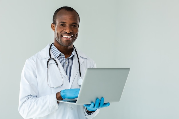 Allegro medico professionista che tiene il suo laptop mentre levandosi in piedi contro il muro bianco
