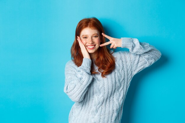 Allegro modello femminile rossa che trasmette buone vibrazioni, sorridente e mostrando segno di pace, in piedi su sfondo blu.