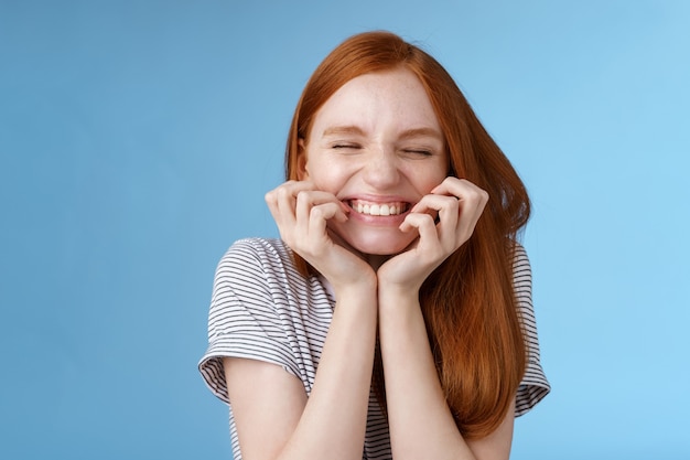 Allegro ridacchiando spensierato zenzero ragazza sguardo felice luminoso chiudere gli occhi sorridenti contenti tenere le mani guance divertirsi sensazione eccitazione gioia trionfante tifo buone notizie, in piedi sfondo blu.