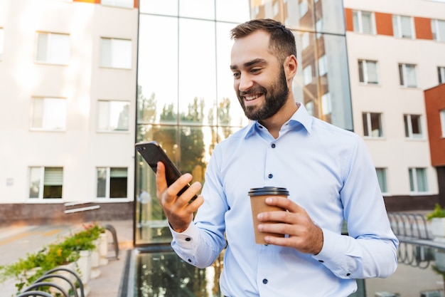 allegro sorridente felice giovane uomo d'affari in piedi vicino al centro affari che tiene tazza di caffè utilizzando il telefono cellulare.