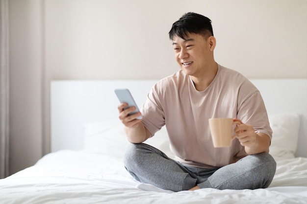 Allegro uomo asiatico rilassato che usa lo smartphone a letto