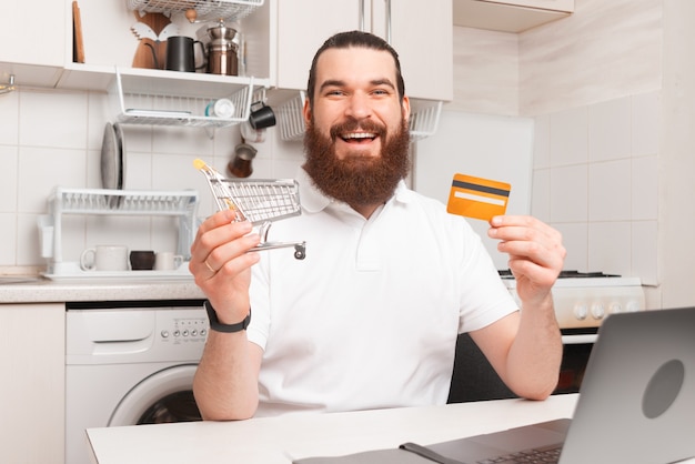 Allegro uomo barbuto sorridente e tenendo il carrello della spesa e la carta di credito davanti al computer portatile