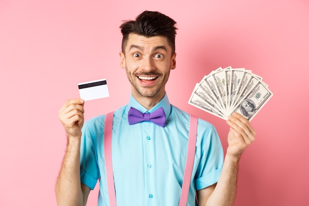 Allegro uomo caucasico con baffi e farfallino che mostra la carta di credito in plastica con soldi in dollari, sorridendo alla telecamera, in piedi su sfondo rosa.