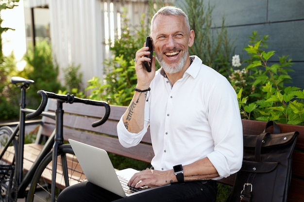 Allegro uomo d'affari di mezza età che sorride parlando al telefono usando il suo computer portatile mentre è seduto sul