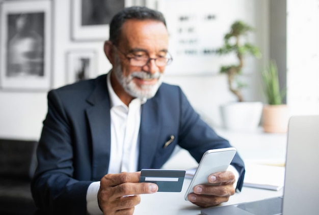 Allegro uomo d'affari europeo senior in occhiali da tuta con laptop usa la carta di credito del telefono al tavolo dentro