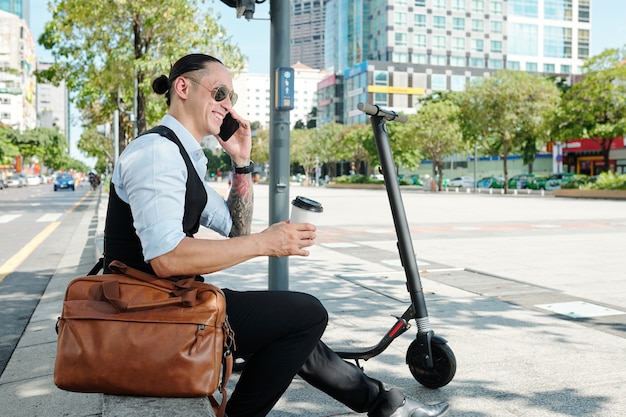 Allegro uomo d'affari positivo seduto all'aperto accanto a uno scooter elettrico, bevendo caffè da asporto e parlando al telefono