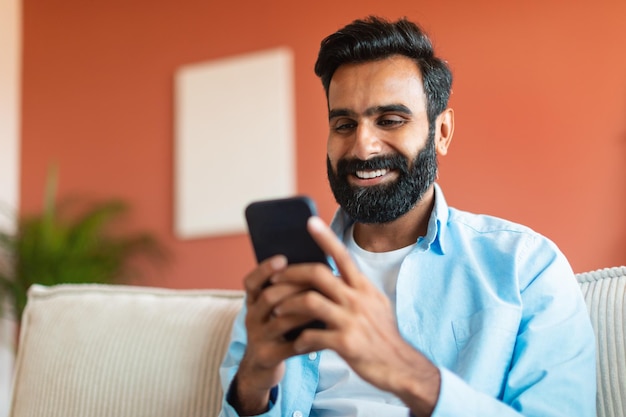 Allegro uomo indiano che utilizza il telefono cellulare navigando in internet seduto al coperto