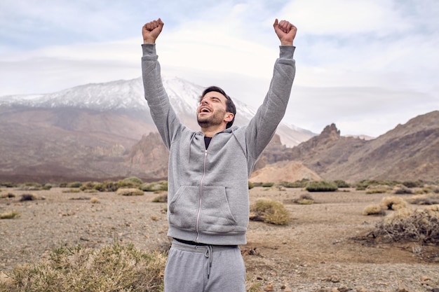 Allegro viaggiatore maschio che alza le braccia mentre celebra il raggiungimento della scalata in montagna durante il viaggio a Tenerife nelle Isole Canarie