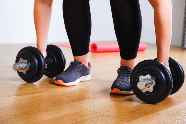 allenamento a casa donna bionda irriconoscibile in leggings neri e scarpe da ginnastica che fa allenamento di forza con manubri su un tappetino rosa a casa bottiglia d'acqua sullo sfondo