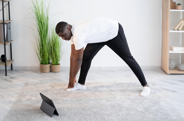 Allenamento a casa Uomo nero felice Lezione online Godersi il fitness Ragazzo africano concentrato che si piega in avanti facendo un allenamento di stretching cercando l'interno della stanza luminosa del computer portatile