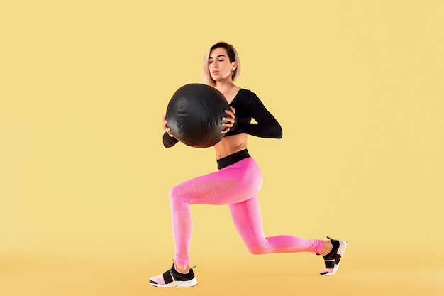 Allenamento donna forte con palla medica. Foto della donna latina sportiva in abiti sportivi alla moda sulla parete gialla. Forza e motivazione.