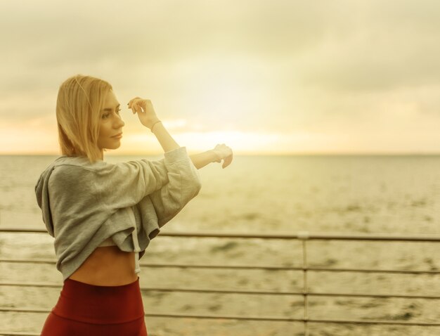 Allenamento mattutino. Concetto di stile di vita sano. La giovane donna attraente in abiti sportivi allunga la mano prima dell'esercizio sulla spiaggia all'alba. Riscaldamento