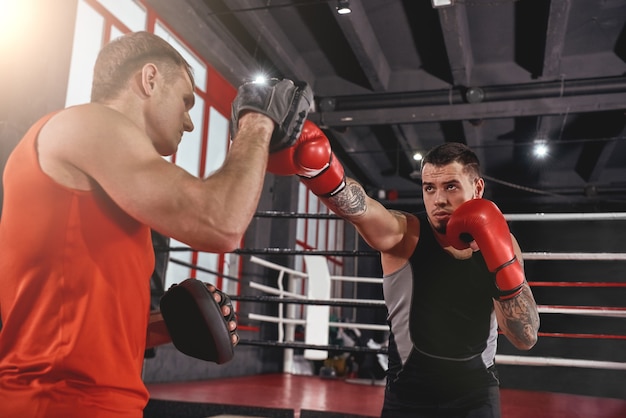 Allenamento per il grande giorno. Uomo muscoloso atletico tatuato in abbigliamento sportivo allenamento su zampe da boxe con partner in palestra di boxe nera