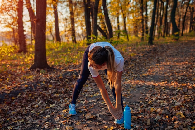 Allenandosi ed esercitandosi nel parco di autunno. Donna che allunga le gambe chinandosi all'aperto. Stile di vita sano attivo