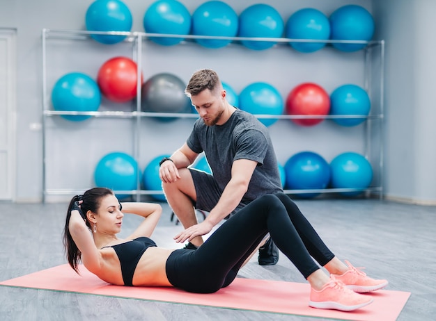 Allenatore che aiuta la giovane donna a fare esercizi addominali sullo sfondo di palle fitness Bella donna che fa addominali su una stuoia e un uomo la aiuta in palestra