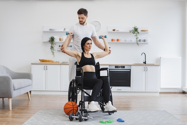 Allenatore che aiuta una donna matura in sedia a rotelle a sollevare le braccia con i manubri