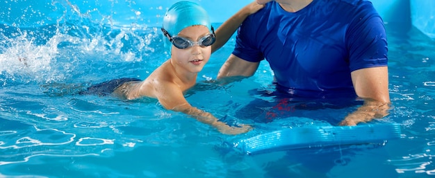 Allenatore maschio che insegna al ragazzo in età prescolare come nuotare nella piscina coperta con la tavola flutter della piscina