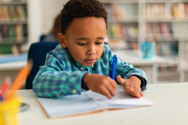 Allievo afroamericano che impara a scrivere nel quaderno con la penna seduto alla scrivania nella scuola primaria