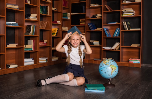 Allievo della ragazza felice in uniforme scolastica che si siede con un libro sulla sua testa e che ride in classe