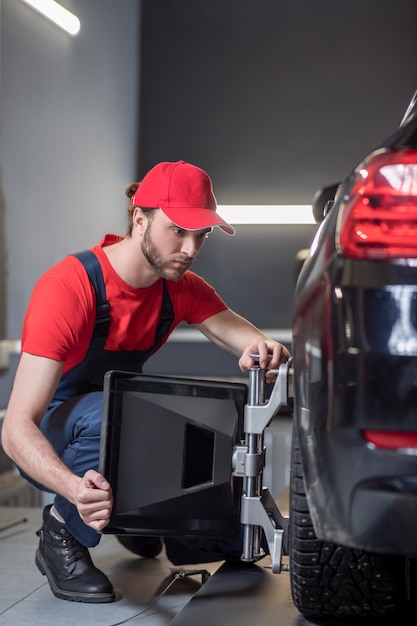 Allineamento delle ruote. Giovane uomo adulto concentrato in tuta accovacciato vicino alla ruota anteriore con un dispositivo speciale in officina