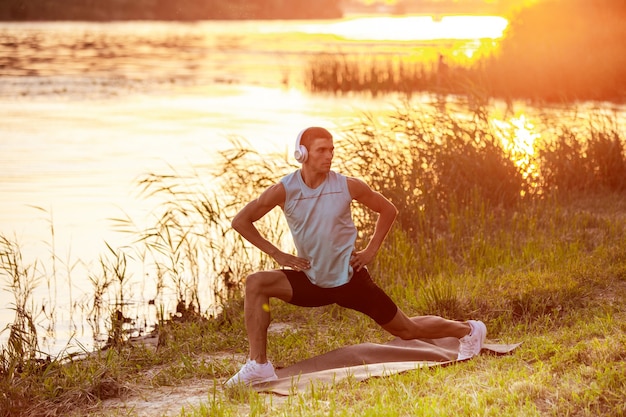 Allungamento. Un giovane uomo atletico che si allena, si allena ascoltando musica in riva al fiume all'aperto. Concetto di stile di vita sano, benessere, sport, attività, perdita di peso. Ispirato dalla natura, l'estate.