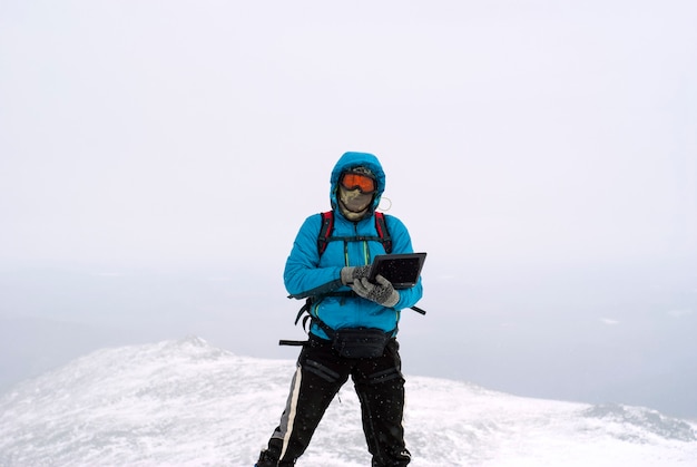 Alpinista che lavora con un laptop in inverno in cima a una montagna durante la bufera di neve