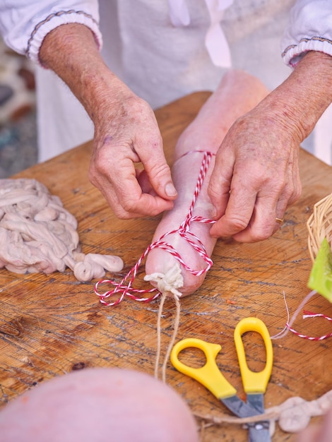 Alto angolo di raccolto anonimo persona matura in uniforme sterile che lega salsiccia cruda appena riempita con filo in piedi al tavolo di legno in macelleria