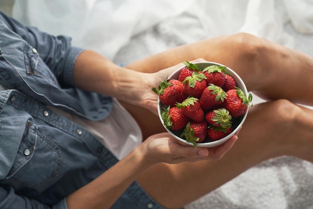 Alto angolo di raccolto femminile che tiene una ciotola in ceramica con fragole appetitose mentre è seduta su tessuto bianco in una stanza luminosa