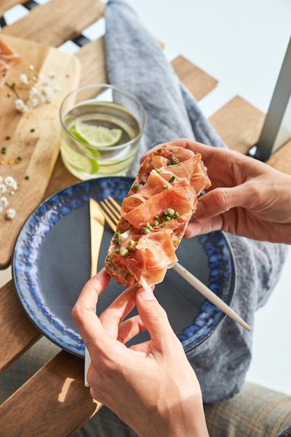 Alto angolo di raccolto femminile senza volto che mangia deliziosi toast con prosciutto e capperi seduti al tavolo di legno con bicchiere di acqua di lime e piatto di ceramica sulla terrazza del ristorante