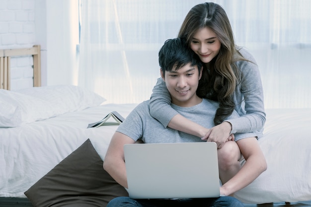 Amante o coppie asiatici felici del primo piano che parlano e che sorridono quando utilizzano il computer portatile di tecnologia sul letto in b