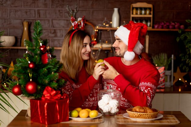 Amanti felici un uomo con una ragazza in maglioni rossi in cucina con un albero di Natale a casa festeggiano il nuovo anno o il Natale gioendo, abbracciando e baciando