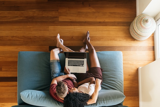 Amanti multietnici che si siedono insieme sullo strato con il computer portatile