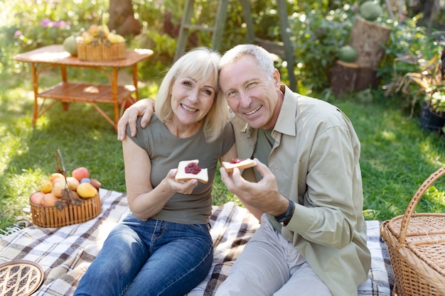 Amare i coniugi anziani che fanno picnic seduti su una coperta in giardino e mangiano toast con marmellata sorridente