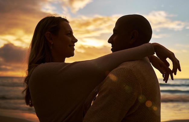 Amare la giovane coppia interrazziale affettuosa in silhouette di fronte all'altro mentre trascorrono del tempo insieme sulla spiaggia al tramonto Gli amanti condividono un momento romantico contro il cielo dorato mentre sono in vacanza