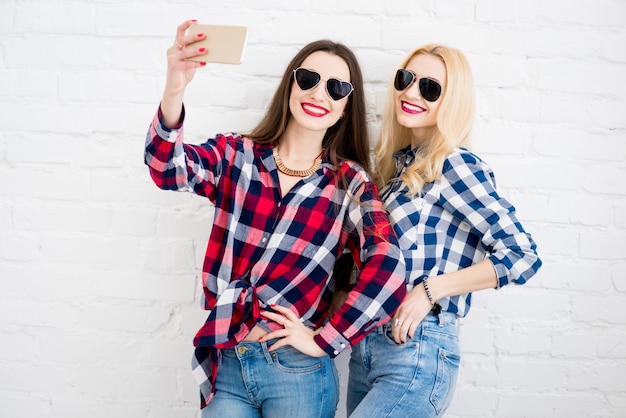 Amiche in camicie a scacchi che fanno selfie ritratto con il telefono sullo sfondo del muro bianco