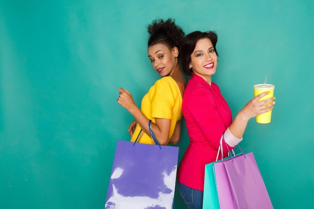 Amiche multietniche felici con borse della spesa e bevande da asporto. Due entusiasti dello shopping a sfondo turchese sudio con copia spazio