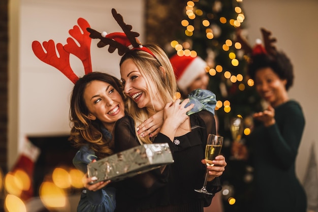 Amiche sorridenti che si abbracciano e si scambiano regali mentre si riuniscono a casa per festeggiare Natale o Capodanno.
