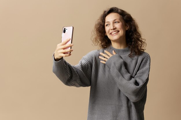 Amichevole bella donna riccia in maglione casual grigio che fa videochiamata con il telefono in posa isolato su sfondo beige pastello Concetto di comunicazione a distanza della rete di social media Spazio di copia