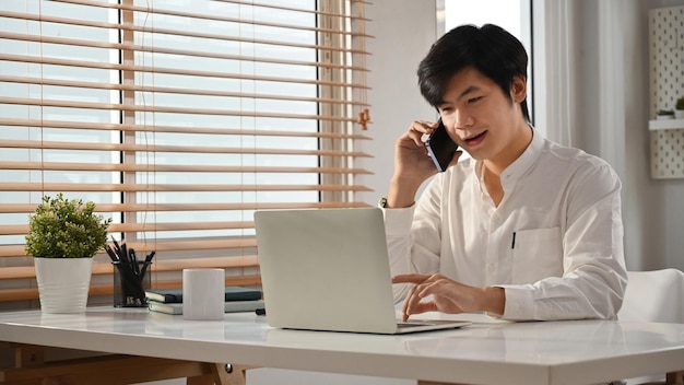 Amichevole consulente finanziario maschile che consulta il cliente a distanza tramite telefonata sul cellulare e utilizzando il laptop sul posto di lavoro moderno