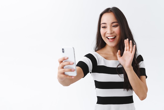 Amichevole donna asiatica in uscita che si fa selfie o parla tramite l'app del telefono videochiamata dì ciao connettiti al live streaming sorridente saluto follower in piedi sfondo bianco registra vlog