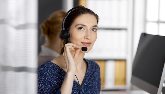 Amichevole donna d'affari asiatica o latinoamericana che parla con l'auricolare in ufficio. Call center e gruppo di persone diverse nel mondo degli affari.