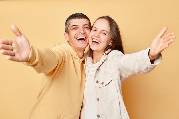 Amichevole donna sorridente e uomo che indossa abiti in stile casual in piedi isolato su sfondo beige che allarga le mani invitando mostrando gesto di benvenuto ridendo di felicità