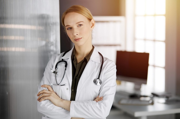 Amichevole dottoressa sorridente in piedi nella clinica soleggiata. Ritratto di donna medico amichevole. Concetto di medicina.