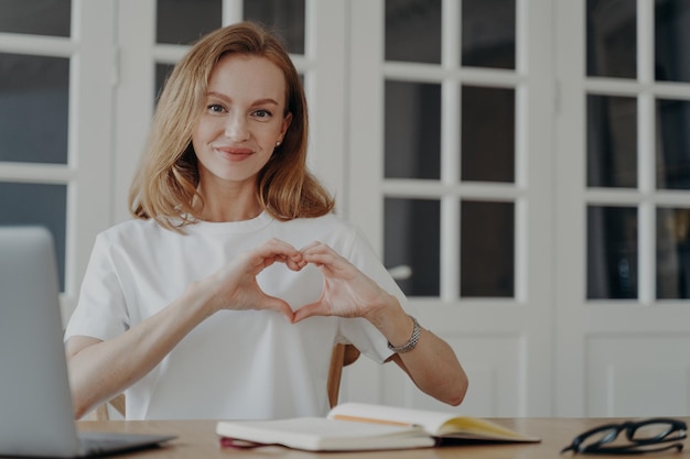 Amichevole femmina riconoscente mostra il gesto della mano del cuore seduto al computer portatile sul posto di lavoro Feedback positivo