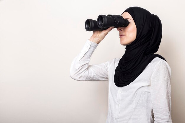 Amichevole giovane donna musulmana in camicia bianca e hijab girata di lato guarda attraverso il binocolo Banner