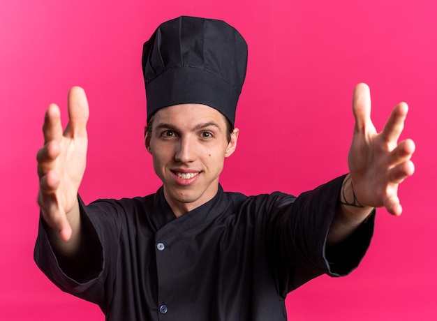 Amichevole giovane maschio biondo cuoco in uniforme da chef e berretto guardando la telecamera facendo gesto di benvenuto isolato su parete rosa