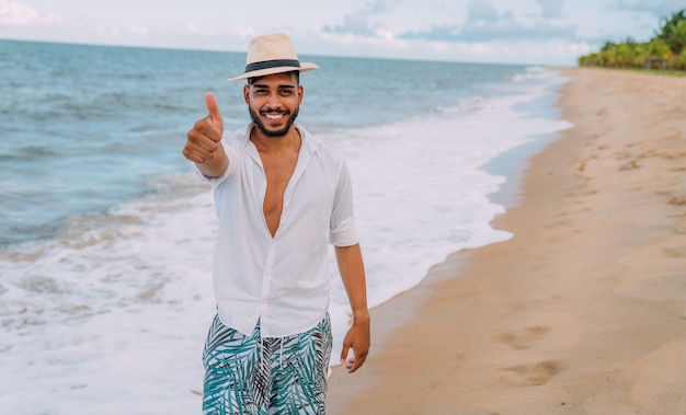 Amichevole giovane uomo latino-americano che fa il gesto del pollice in su sulla spiaggia, con indosso un cappello