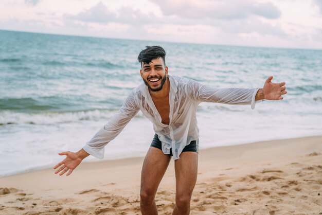 Amichevole giovane uomo latinoamericano fiducioso e sorridente con le braccia aperte che guarda l'obbiettivo sulla spiaggia