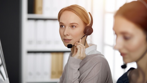 Amichevole imprenditrice caucasica parlando con l'auricolare in ufficio soleggiato. Call center e gruppo di persone diverse nel mondo degli affari.